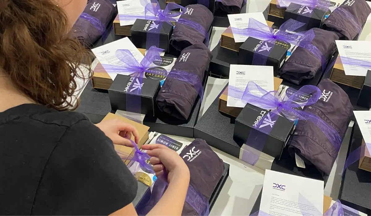 A woman in a black shirt hand-assembles rows of DXC-branded gift boxes, each containing items wrapped in purple tulle with ribbon, a printed card, and a copy of Simon Sinek's book.