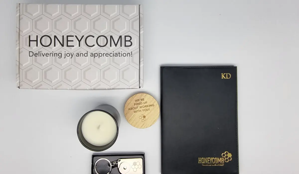 A Honeycomb-branded white gift box reading "Delivering joy and appreciation," arranged on a light surface alongside a black hardcover notebook, a scented candle, and a keychain.