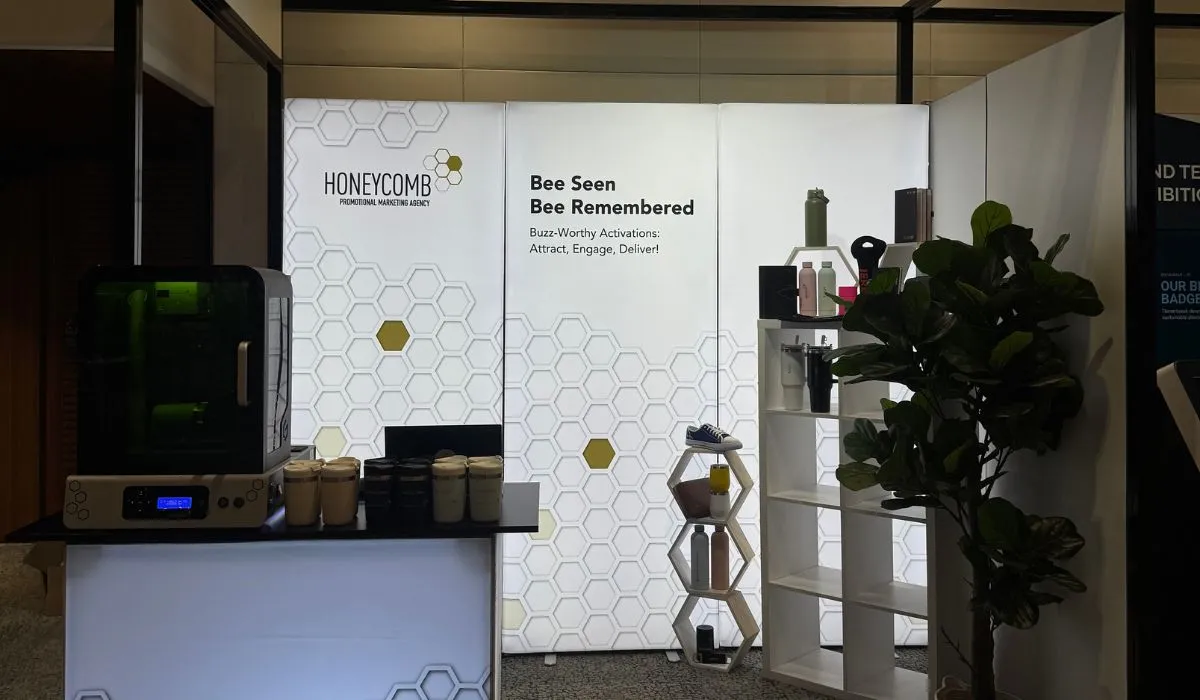 Honeycomb Agency trade show booth with a laser engraving machine, branded drink bottles on display shelves, and a "Bee Seen, Bee Remembered" backdrop.