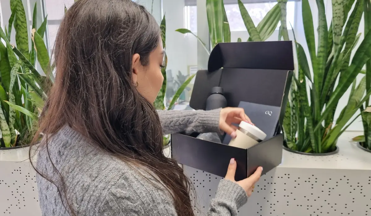 woman with long brown hair opens a black spring corporate gift box, lifting out a tissue-wrapped item, with green potted plants in the background.
