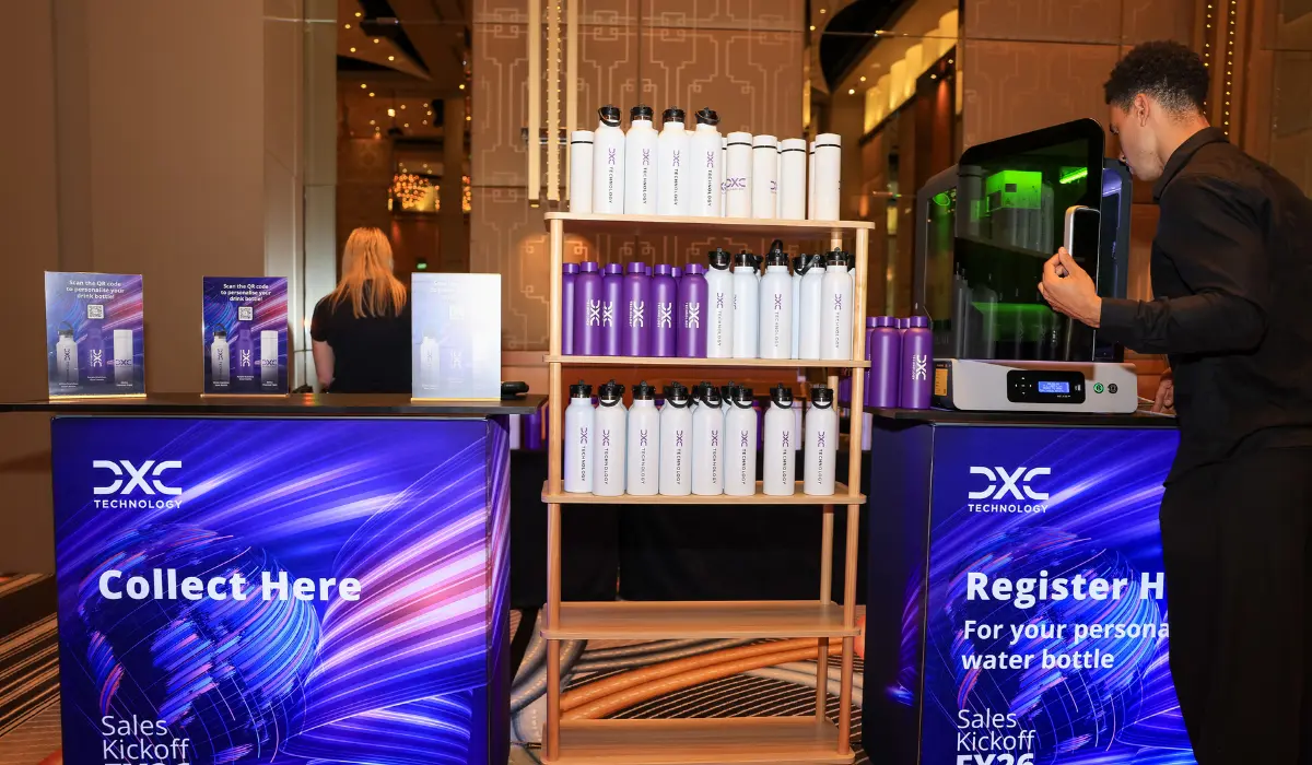 An activation stand with "Collect Here" and "Register" screens, shelves of personalised bottles, and staff serving guests