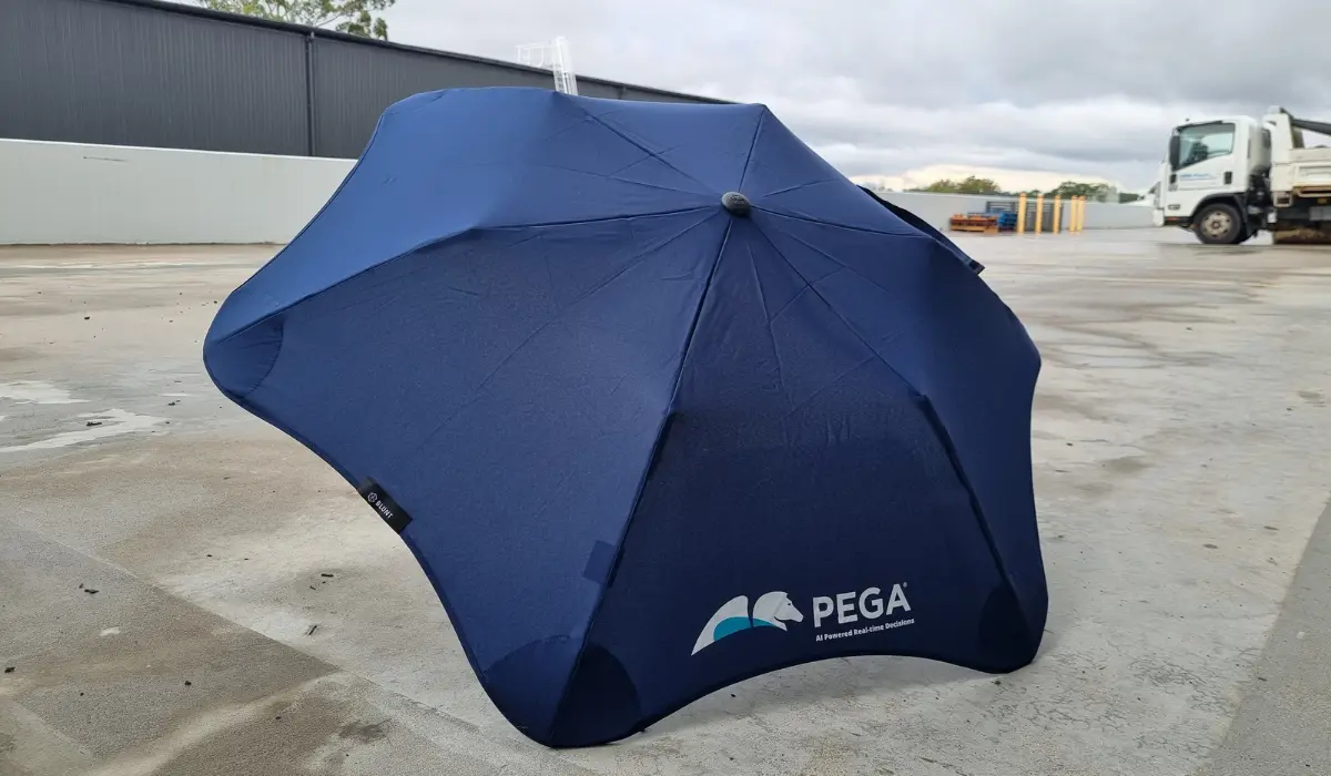 A navy blue umbrella printed with the PEGA logo, set up outdoors on a paved lot with a truck in the background.