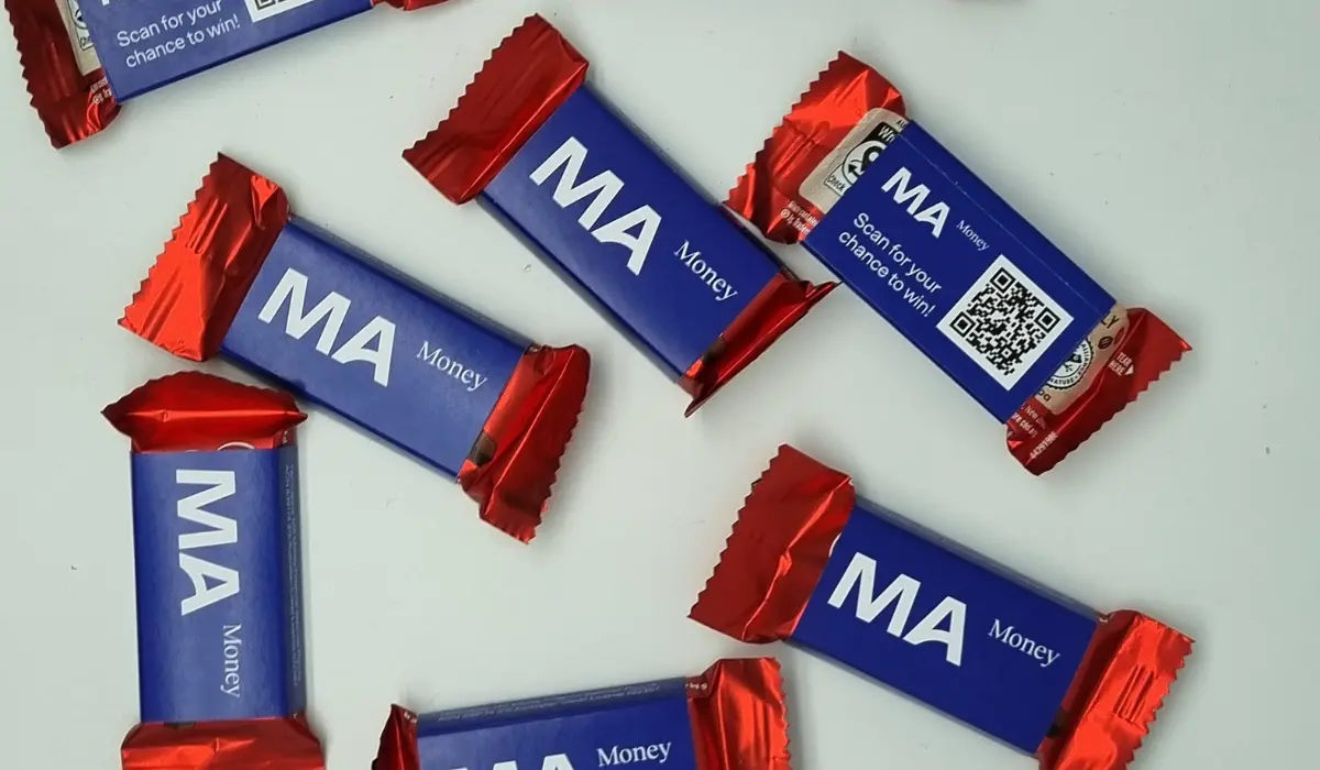 Several custom branded chocolate bars in blue and red foil wrappers featuring the MA Money logo and a QR code, scattered on a light background.