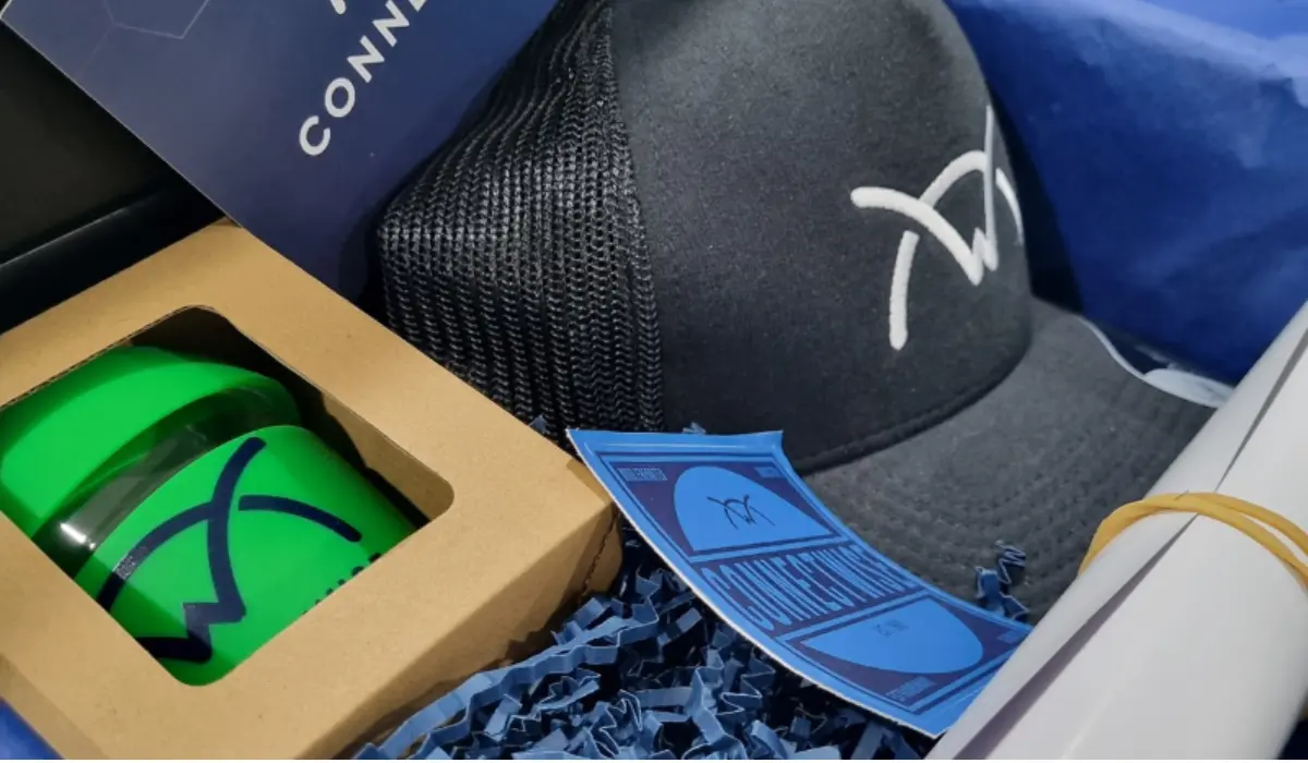 Corporate gift ideas for clients featuring a branded trucker cap, logo tumbler, and card in a gift box with blue crinkle paper.